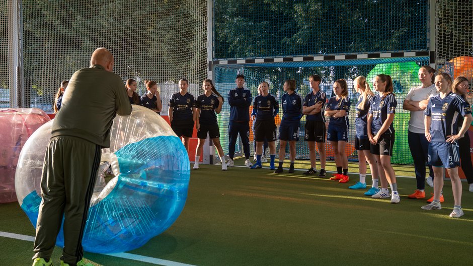 "Teamaktivität im Bubble Soccer: Vorfreude auf das Spiel" Gruppe von Personen auf einem Indoor-Fußballfeld, umgeben von Zorbing-Bällen, hören einer stehenden Person zu.