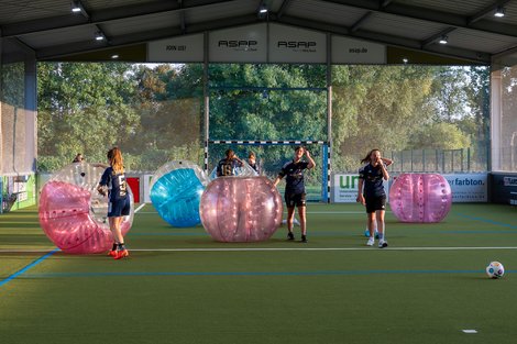 "Spiel und Spaß im Bubble-Soccer-Parcours" Personen spielen Bubble-Fußball in einer Halle, umgeben von großen, farbigen Plastikkugeln auf einem Kunstrasenfeld.