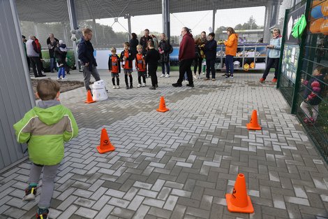 Junge läuft auf einen Parcours zu, während Zuschauer und Kinder in Fußballtrikots neben einem Zaun stehen.