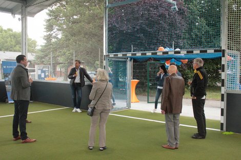 Gruppe von Menschen steht im überdachten Sportbereich. Zwei sprechen, andere hören zu. Ballons im Hintergrund.