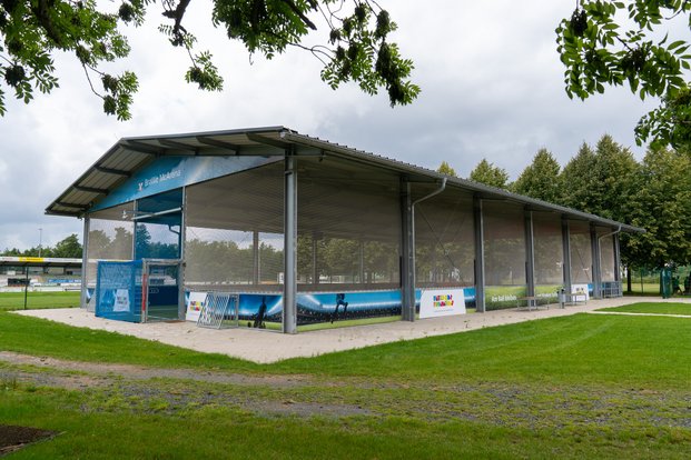 Überdachtes Sportkäfigfeld mit Werbebannern, umgeben von grünem Rasen und Bäumen.