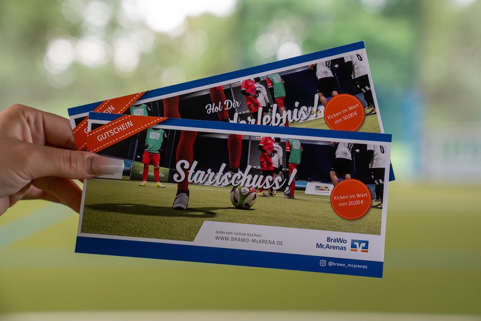 Das Bild zeigt zwei Gutscheine für die BraWo McArenas, die von einer Hand gehalten werden. Auf den Gutscheinen ist ein Bild von einem Fußballspiel abgebildet. Die Beschriftung "Hol Dir das Erlebnis!" und "Startschuss!" ist groß sichtbar. Auf den Gutscheinen sind orangefarbene Kreise mit Informationen über den Wert des Kickens von 20,00 € und 50,00 €. Unten auf den Gutscheinen steht eine Webseite zur Buchung und das Logo der BraWo McArenas.