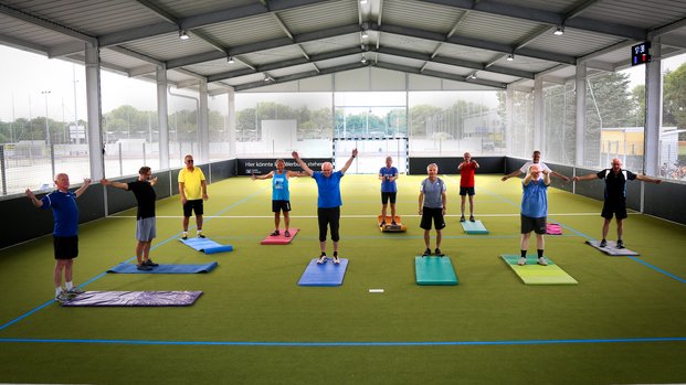 "Gemeinschaftliches Fitness-Training in der Halle" Gruppe von Männern macht Gymnastikübungen auf einem überdachten Sportplatz, alle stehen auf Matten.
