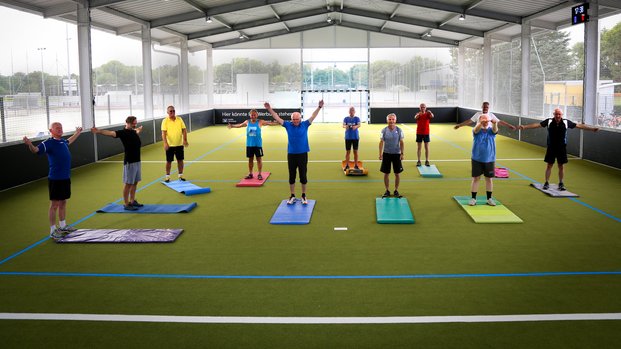 "Gemeinschaftliches Fitness-Training in der Halle" Gruppe von Männern macht Gymnastikübungen auf einem überdachten Sportplatz, alle stehen auf Matten.