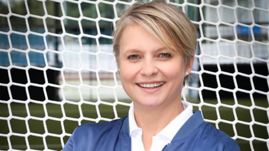 Person vor einem Fußballtor lächelt in die Kamera, trägt eine blaue Jacke und ein weißes Hemd.
