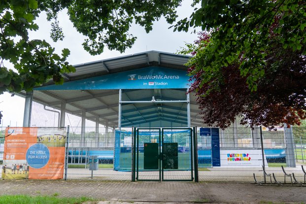 Überdachte Sporthalle mit Netz, Bäumen im Vordergrund und Schildern „BraWo McArena“ und „Die Halle für Alle(s)“.