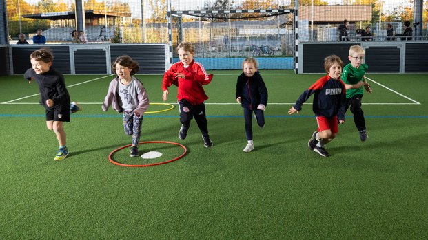 "Fröhlicher Wettlauf: Kinder in Bewegung auf dem Sportplatz" Kinder rennen fröhlich auf einem Kunstrasenfeld. Im Hintergrund sind Netze und Zuschauer zu sehen.