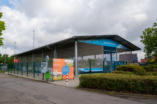 Eine überdachte Sporthalle mit dem Namen "BraWo McArena" ist von einem Zaun umgeben. Ein Werbebanner an der Vorderseite wirbt für die Halle mit der Aufschrift "Die Halle für alle!" Es stehen Informations- und Anmeldehinweise auf dem Banner. Vor der Halle befindet sich eine gepflasterte Fläche mit einem Mülleimer und einem Verbotsschild für die Einfahrt. Im Hintergrund sind einige Bäume sowie ein Gebäude mit Solarpanelen auf dem Dach zu sehen. Wolken bedecken den Himmel.