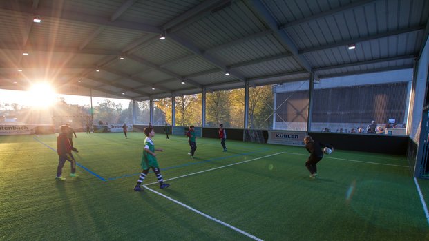 "Abendlicher Jugendfußball: Spielspaß in der Halle im Sonnenuntergang" Kinder spielen bei Sonnenuntergang auf einem überdachten Fußballfeld.