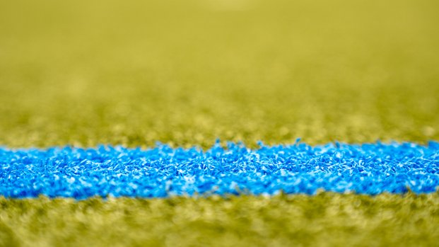 "Spielbeginn: Die blaue Linie auf dem Spielfeld" Nahaufnahme einer blauen Linie auf einem grünen Kunstrasenplatz. Die Linie ist im Mittelpunkt, der Hintergrund ist unscharf.
