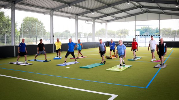 "Gruppendynamik: Outdoor-Fitness" Eine Gruppe von Personen macht Dehnübungen in einer überdachten Sporthalle auf grünen Matten.