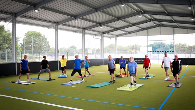 "Gruppendynamik: Outdoor-Fitness" Eine Gruppe von Personen macht Dehnübungen in einer überdachten Sporthalle auf grünen Matten.