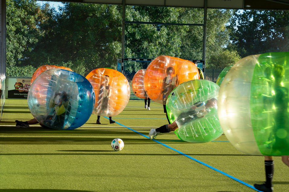 Spieler in aufblasbaren Kugeln spielen auf einem Spielfeld mit einem Fußball, umgeben von Bäumen und einem Netz.