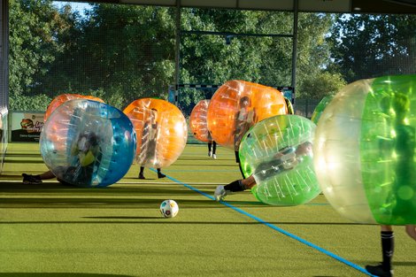 "Bubble-Fußball: Spaß und Action auf dem Spielfeld" Spieler in aufblasbaren Kugeln spielen auf einem Spielfeld mit einem Fußball, umgeben von Bäumen und einem Netz.