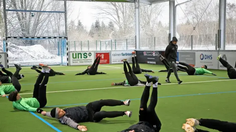 "Intensives Training: Fußballmannschaft bei der Wintervorbereitung" Personen in Sportkleidung machen Gymnastikübungen auf einem Kunstrasenplatz in einer Halle, während ein Trainer beobachtet.