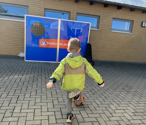 Ein Kind spielt mit einem Fußball vor einer Wand mit Loch auf einem gepflasterten Platz bei sonnigem Wetter.