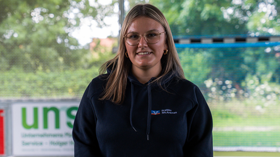 Person in dunklem Hoodie mit Brille, lächelt nach vorne. Im Hintergrund sind Bäume und ein unscharfer Text zu sehen.