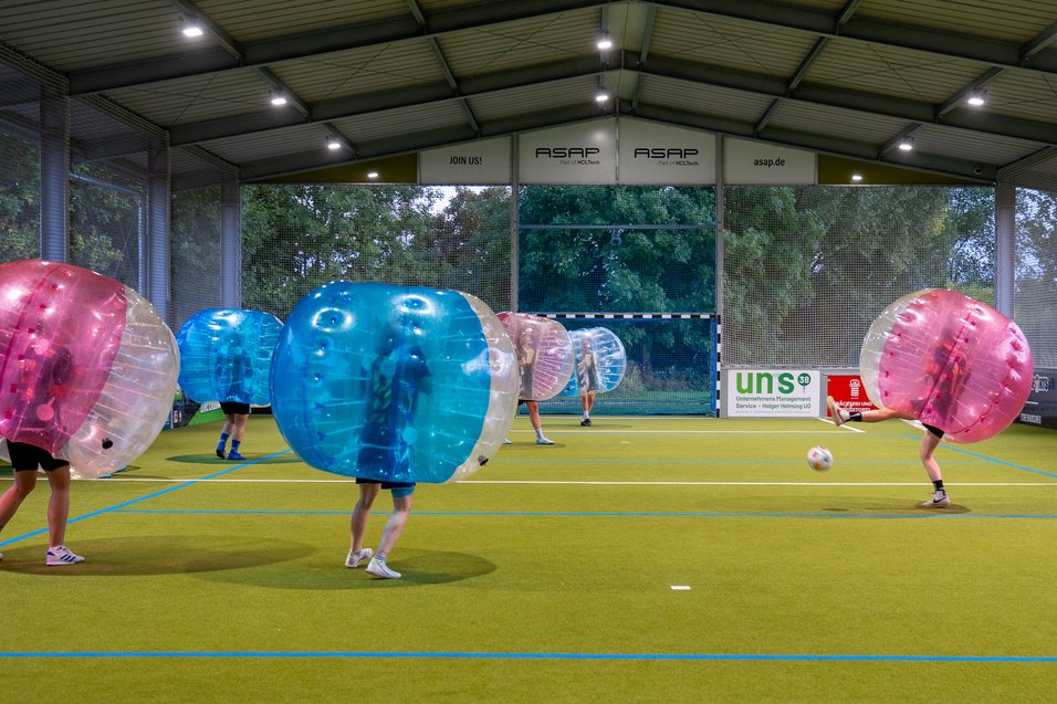 Personen in aufblasbaren Bällen spielen Bubble-Fußball in einer Halle auf Kunstrasen.